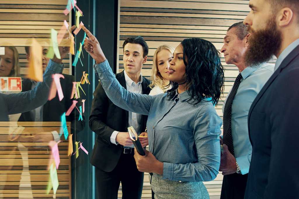 A group discussion in an office workspace uses sticky notes on a glass wall to organize information.