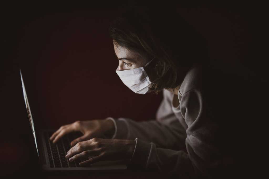 A laptop user wearing a face mask works in a darkened space.