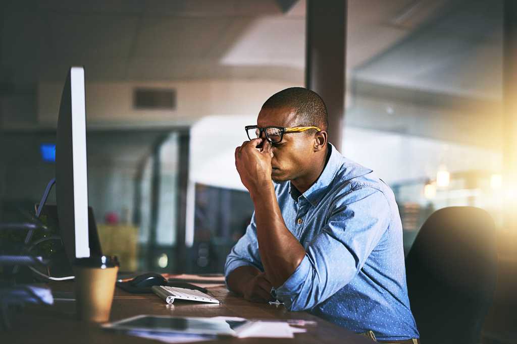 A man looks displeased/stressed/frustrated and pinches his brow while using a computer at work.