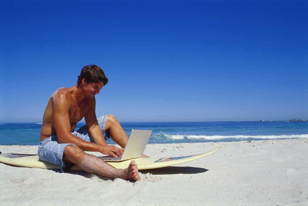Man sitting on surfboard on beach with laptop