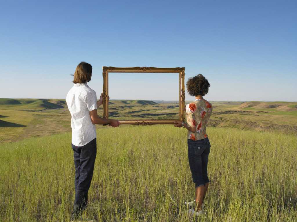 young man and woman standing in field holding empty gold frame