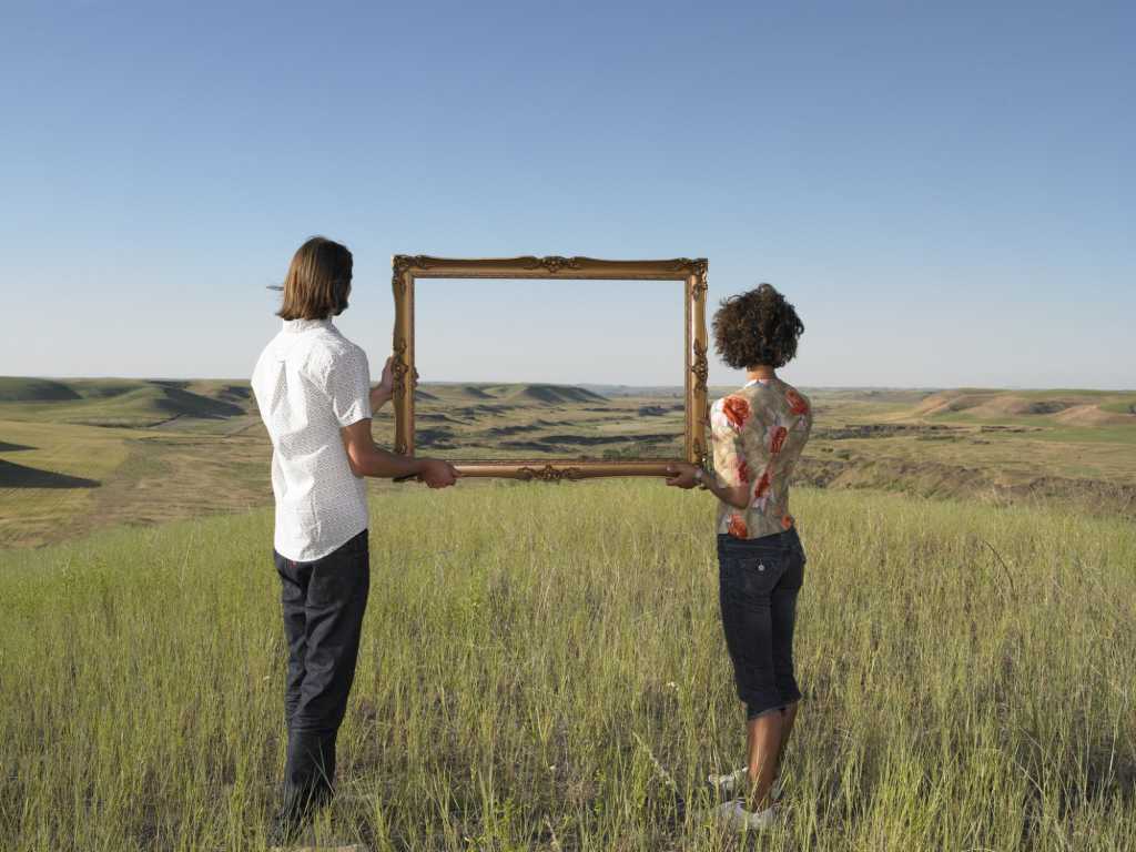 two millennials standing in field holding picture frame against blue sky
