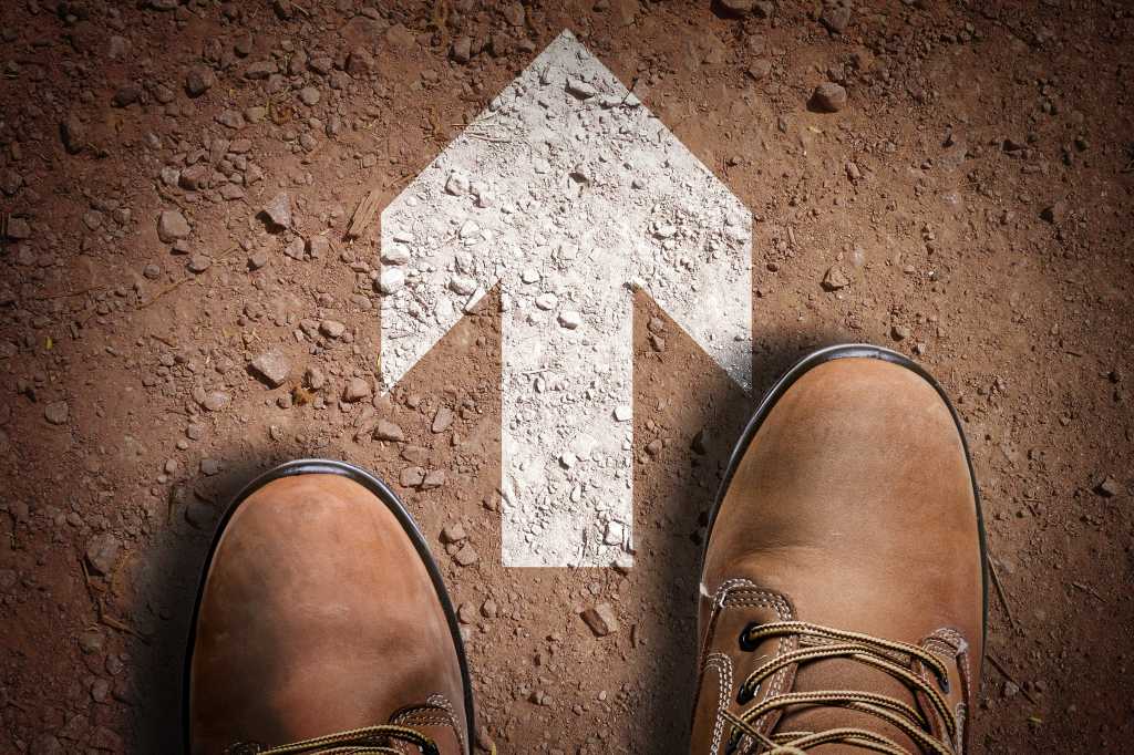 Aerial view of work boots facing a directional arrow on asphalt [forward / progress / future / what