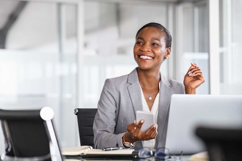 Black business woman using smartphone while working on laptop at office. Smiling mature african american businesswoman looking up while working on phone. Successful woman entrepreneur.