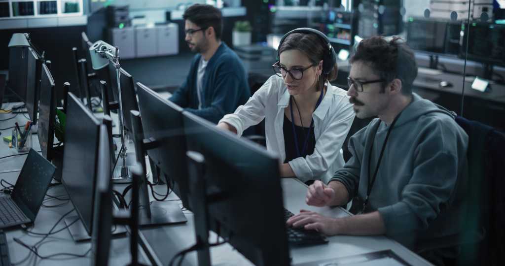 Young Team of Specialists Working on Desktop Computers and Having a Conversation at a Workplace. Female and Male Software Developers Discussing a Solution for Their Artificial Intelligence Project
