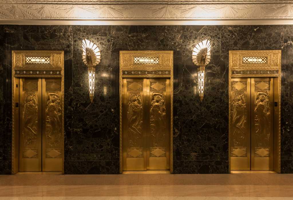 Dramatic golden elevators in fancy urban skyscraper building with relief sculptures and marble wall with lights and sconces