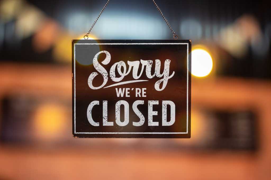 Sorry we're closed sign hanging inside a glass door, colored lights in the background.