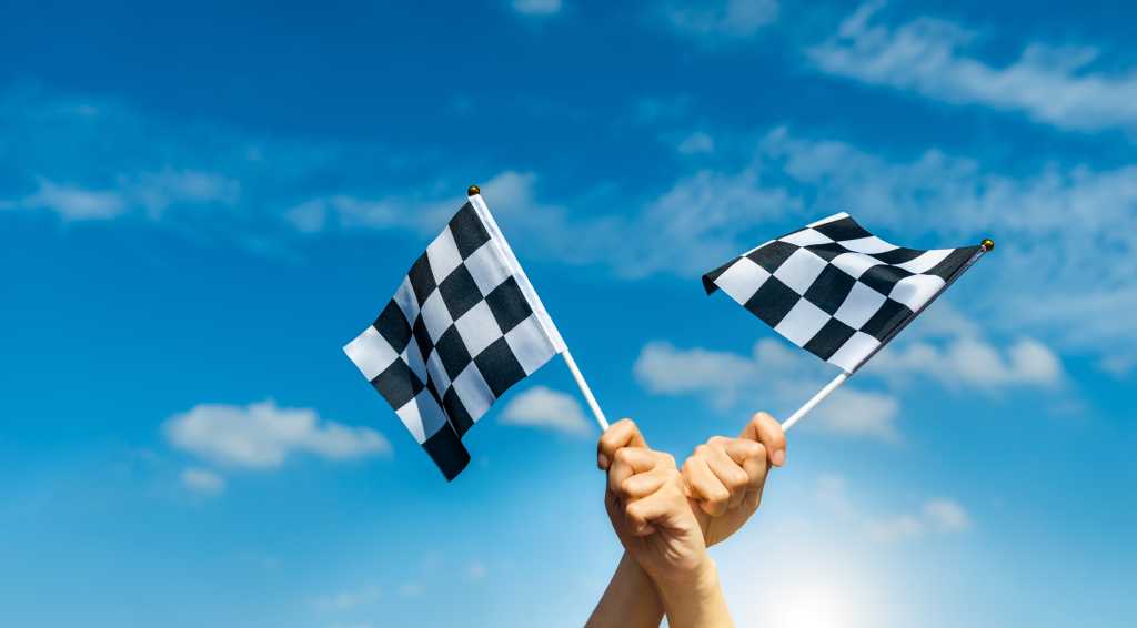 Two checkered flags against a blue sky, held by crossed hands