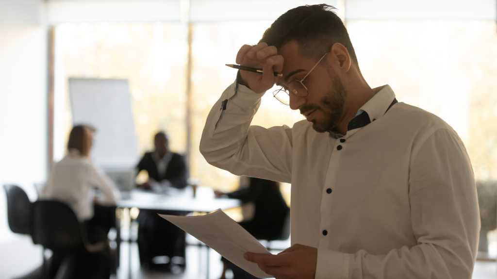 man reading a document and rubbing his forehead indicating stress worry concern with people meeting around a table in the background