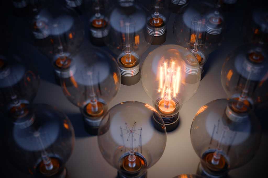 A glowing light bulb stands out from a crowd of unlit bulbs. [ideas / innovation / transformation]