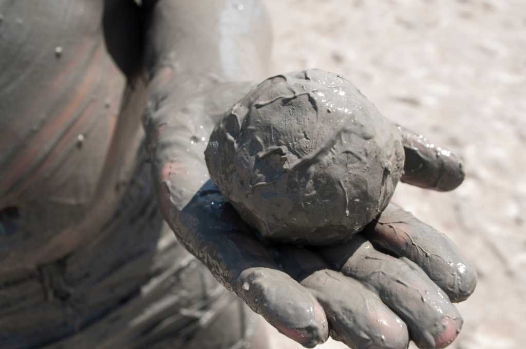 A hand covered in mud presents a ball of mud with mud in the background