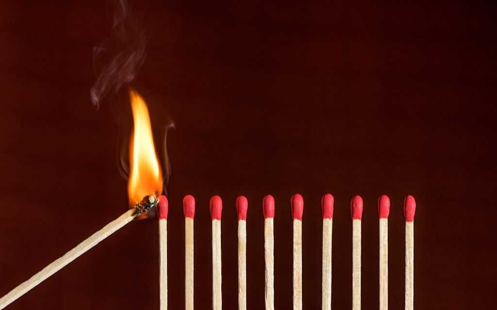 A lit match lights the first in a row of unlit matches. Lit match next to a row of unlit matches. Red phosphorus matches on dark red matte background.