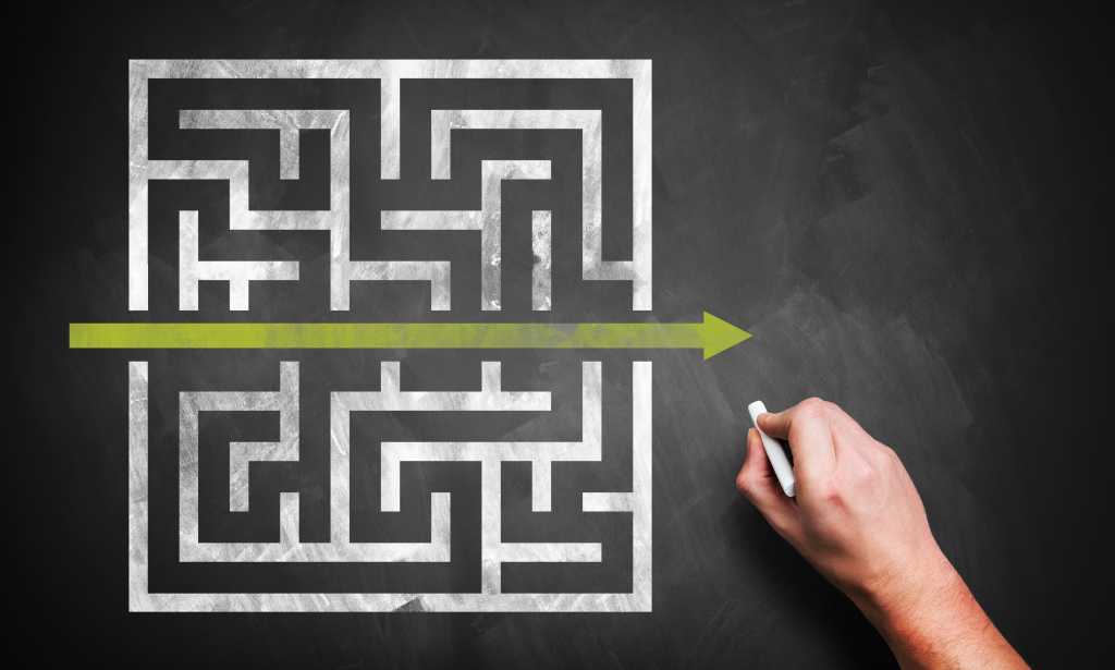 Hand of someone solving a maze drawn in white on a black chalkboard.