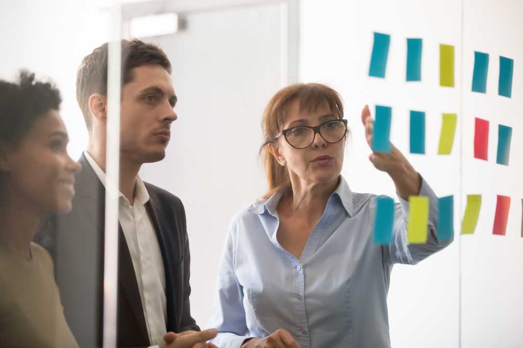 Middle aged businesswoman mentor coach leader explaining project strategy showing post it sticky notes on glass wall, serious business team brainstorm work plan at corporate office stand up meeting