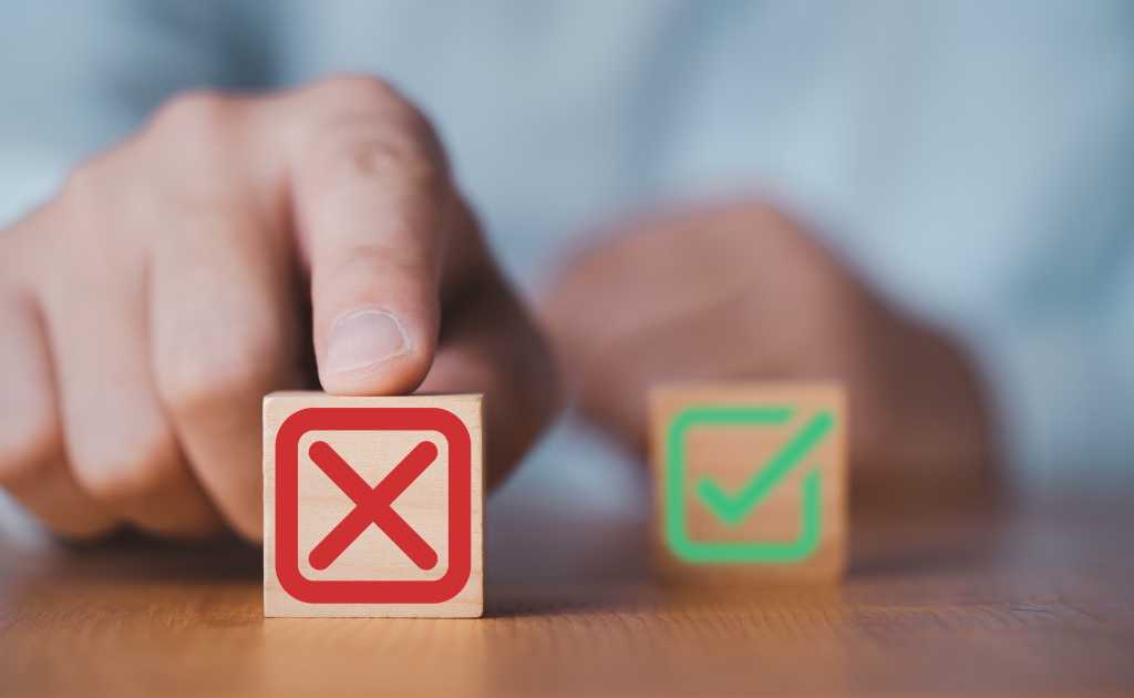 Two building blocks pictured, one with a green check mark and the other with a red x, standing for cancelation. The x is pushed to the foreground by a human hand.