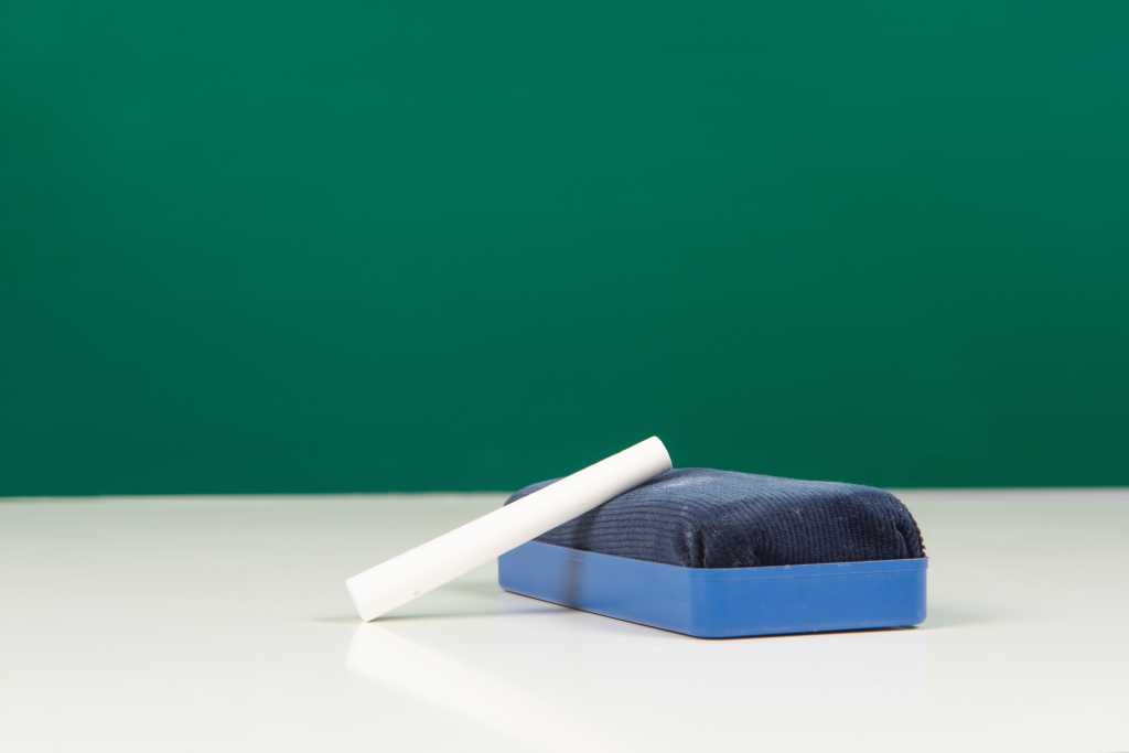 An eraser and chalk sit on a desk in front of a clean, empty blackboard.