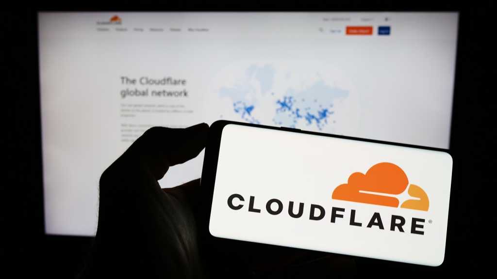 A photograph of a person holding a smartphone with the Cloudflare logo displayed. Behind the phone is a blurred monitor displaying the Cloudflare website.,