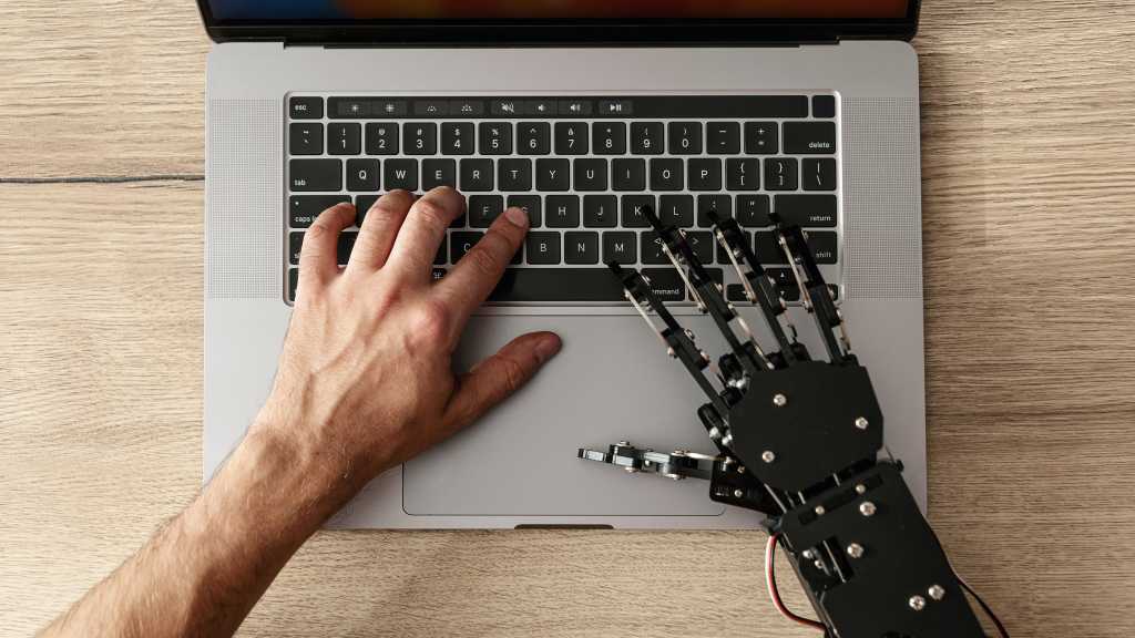 image of a human and robot hand typing at a laptop